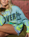 A woman sits on a bench in a rustic room wearing an oversized light blue and neon yellow-green graphic sweatshirt with 'OVER' printed across the front, paired with denim shorts and black knee-high boots. A tiger figurine sits beside her, and framed posters are on the wall behind her.